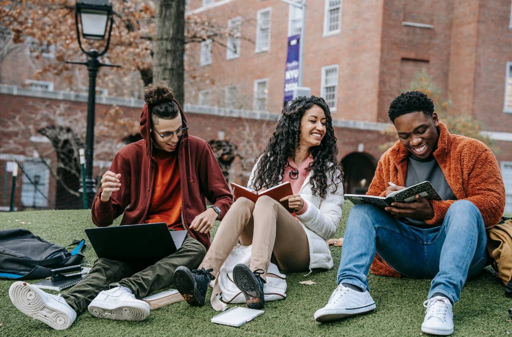 group of 3 students sitting on the grass laughing and looking at tablets and books